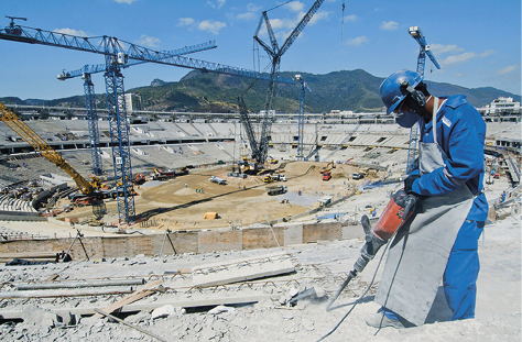 Imagem: Fotografia. Um homem uniformizado com macacão azul, capacete, máscara na região da boca e nariz, protetor auditivo, jaleco e luvas manipula um instrumento ligado a um fio e que perfura uma estrutura de concreto. Ao fundo, um estádio em construção com arquibancada montada e muitas gruas no espaço.  Fim da imagem.