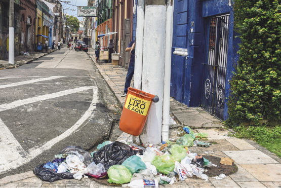 Imagem: Fotografia. Destaque de uma lixeira presa a um poste em uma via. O recipiente está vazio e o chão está cheio de sacos de lixo, alguns estão abertos.  Fim da imagem.