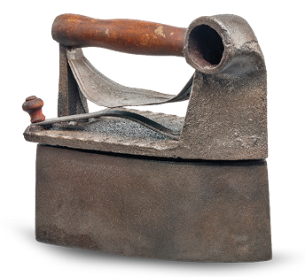 Imagem: Fotografia. Um ferro de passar que apresenta base larga e tampa com rebarba e pegador de madeira com um orifício na lateral.  Fim da imagem.
