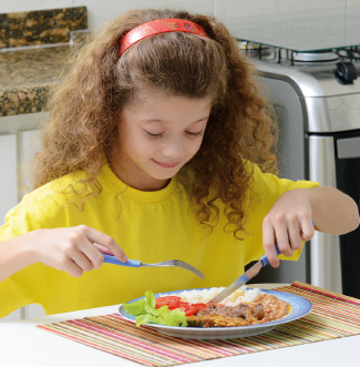 Imagem: Fotografia. Na cozinha, uma menina está sentada à mesa posta com um prato de comida com arroz, feijão, sala e carne. Ela olha a comida sorridente e segura um garfo e faca.  Fim da imagem.