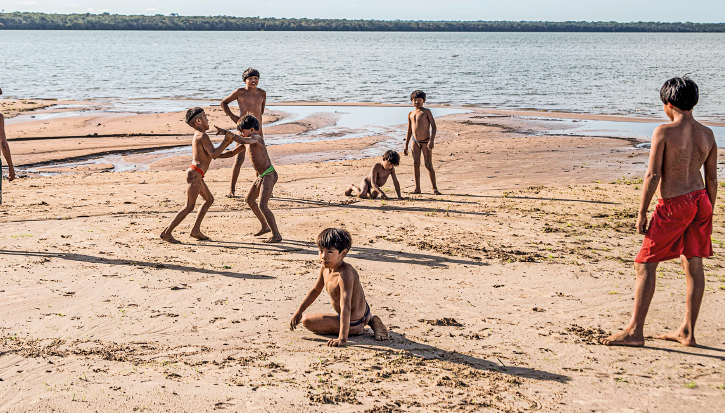 Imagem: Fotografia. Na areia à beira-mar estão sete meninos com características indígenas, como pele parda, olhos puxados e cabelo liso curto e preto. Em primeiro plano, um deles está sentado e olha para trás e ao lado dele um jovem está de pé e olha para frente, onde dois outros garotos brincam juntos de pé, um terceiro observa e há outro sentado mexendo na areia e um de pé ao seu lado. Eles estão sem camisa, usam bermudas ou sungas.  Fim da imagem.