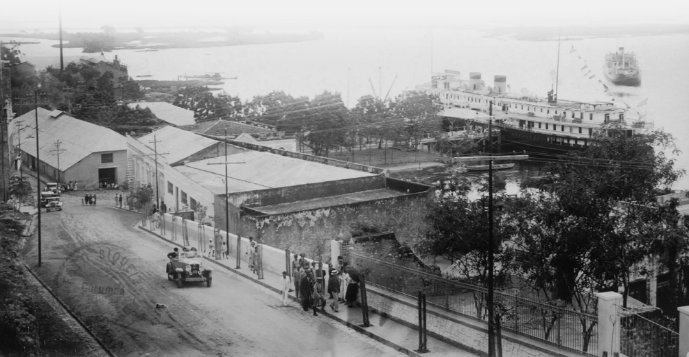 Imagem: Fotografia em preto e branco. Plano aberto de uma via de terra na qual passa um automóvel e há muitas pessoas agrupadas em um trecho da calçada à frente de uma área com muitas árvores e cercada por um portão. Na região, há construções compridas e térreas. Ao fundo, há um terreno com árvores e uma grande embarcação atracada no mar. Fim da imagem.