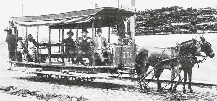 Imagem: Fotografia em preto e branco. Destaque de uma via na qual um pequeno bonde, estrutura coberta, aberta nas laterais e com rodas, é ligado por cordas a dois burros. Dentro do transporte há cerca de seis passageiros, em sua maioria homens, e um condutor uniformizado e de pé.  Fim da imagem.