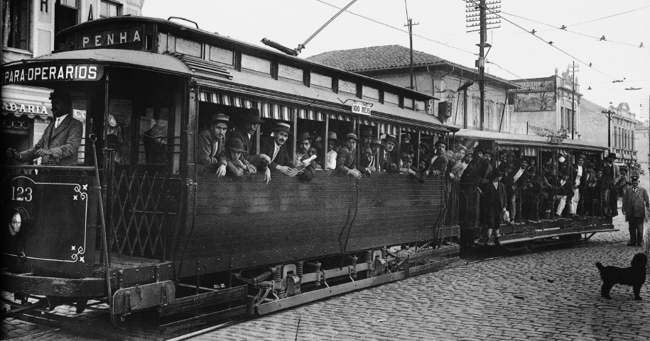 Imagem: Fotografia em preto e branco. Destaque de um bonde longo dividido em três partes. Na primeira, está a cabine com o condutor e acima da qual há um letreiro com a identificação “Penha”. Na segunda, a lateral é coberta e as janelas amplas, nas quais estão muitas pessoas, majoritariamente homens de paletó e chapéu, que posam para foto. A terceira parte é menor, com as laterais descobertas e muitas pessoas aglomeradas de pé. A rua é feita de pedras, há um cachorro no local e fiação na qual é ligada o bonde.  Fim da imagem.