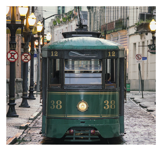 Imagem: Fotografia. Destaque frontal de um bonde verde com farol ao centro da parte dianteira. Ele apresenta o número 38, janelas amplas e é ligado a uma fiação pela parte superior. A rua onde trafega é estreita, feita de pedras e os postes de iluminação tem design antigo e estão acesos. Fim da imagem.