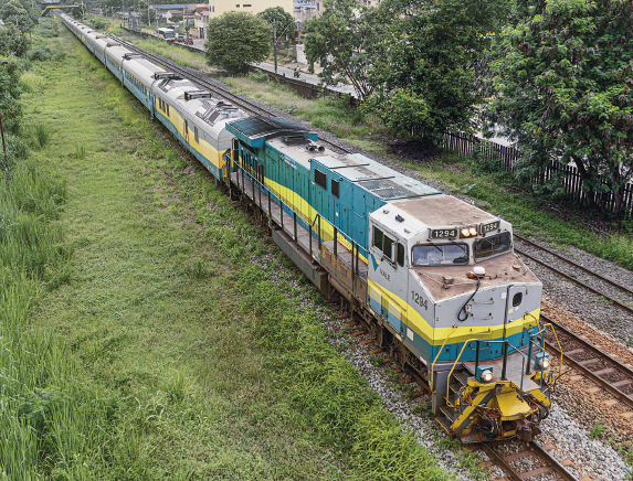 Imagem: Fotografia. Vista aérea de um trem com muitos vagões que se locomove sobre um trilho em meio à área gramada e árvores. A parte frontal apresenta uma cabine com vidros compridos.  Fim da imagem.