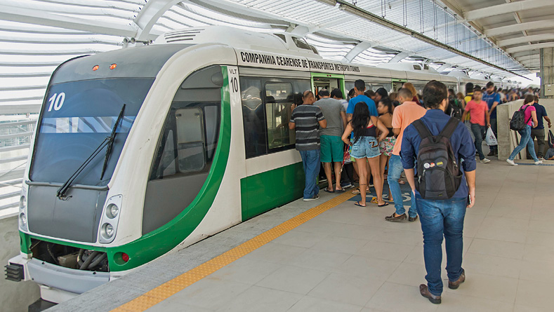 Imagem: Fotografia. Destaque de uma plataforma de metrô coberta onde muitas pessoas aguardam para embarcar no transporte enquanto outras saem em direção à escada.  Fim da imagem.