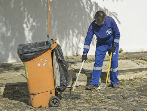 Imagem: Fotografia. Uma mulher uniformizada com macacão azul de manga longa e bota grossa limpa uma via com auxílio de uma vassoura e uma pá. Ao lado, está o carrinho de lixo alto, com um par de rodas, laranja e com saco preto. Fim da imagem.