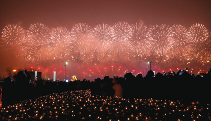 Imagem: Fotografia. Durante a noite, há muitos pontos luminosos no solo e no horizonte há diversos fogos de artifício no céu. Fim da imagem.