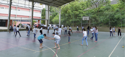 Imagem: Fotografia. Em uma quadra poliesportiva coberta, muitas crianças interagem em pequenos grupos espalhados. Fim da imagem.