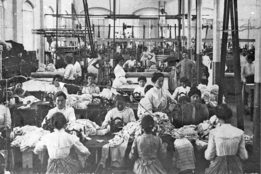 Imagem: Fotografia em preto e branco. No interior de um grande galpão, diversas mulheres trabalham sentadas diante de mesas de com máquinas de costura e muitas roupas. Elas usam coque e longos vestidos.  Fim da imagem.
