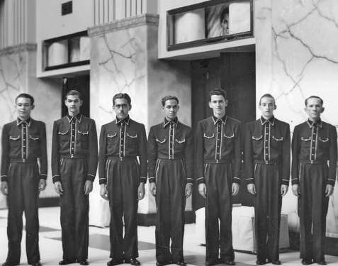 Imagem: Fotografia em preto e branco. Sete homens uniformizados com camisa e calça posam para foto enfileirados lado a lado diante da fachada de um prédio.  Fim da imagem.