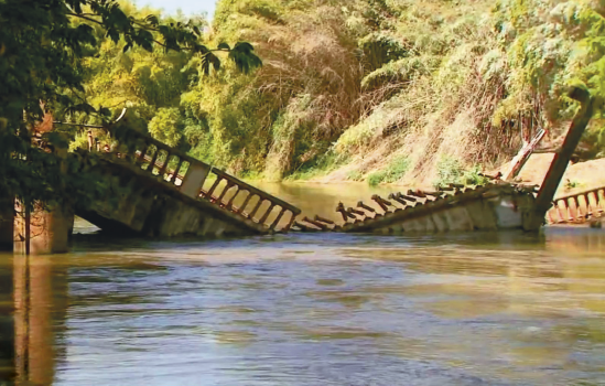 Imagem: Fotografia. Uma ponte quebrada em um rio margeado por vegetação. Fim da imagem.