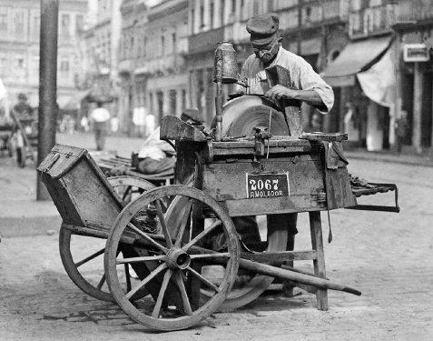 Imagem: Fotografia em preto e branco. Em uma via, um senhor de boina e óculos está de pé ao lado de um carrinho de madeira. Ele segura um instrumento próximo de uma superfície redonda.   Fim da imagem.