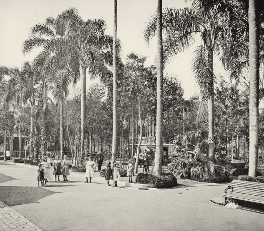 Imagem: Fotografia em sépia. Em um espaço aberto com palmeiras e bancos, algumas pessoas passeiam.  Fim da imagem.