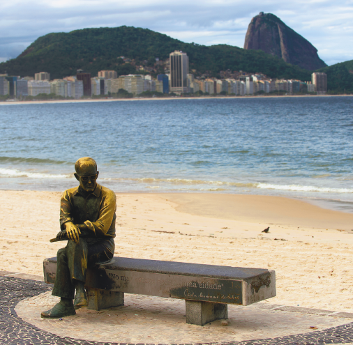 Imagem: Fotografia. Destaque de uma estátua de um homem calvo sentado com as pernas cruzadas em um banco na orla de uma praia. A faixa de areia e o mar estão ao fundo. No horizonte, a cidade com prédios e morros.  Fim da imagem.