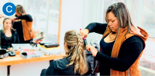 Imagem: C. Fotografia. Uma mulher de pé ajeita o cabelo de uma mulher sentada à sua frente e diante do espelho.  Fim da imagem.
