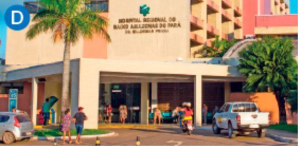 Imagem: D. Fotografia. Fachada de um prédio à frente do qual caminham algumas pessoas e há uma caminhonete estacionada. Próximo à porta de entrada, há alguns bancos onde aguardam algumas pessoas.  Fim da imagem.