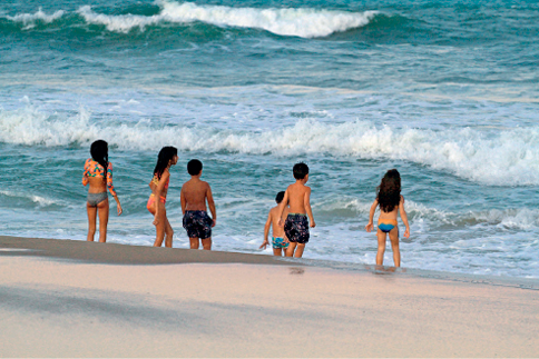 Imagem: Fotografia. Seis crianças com roupa de banho observam o mar da encosta.  Fim da imagem.