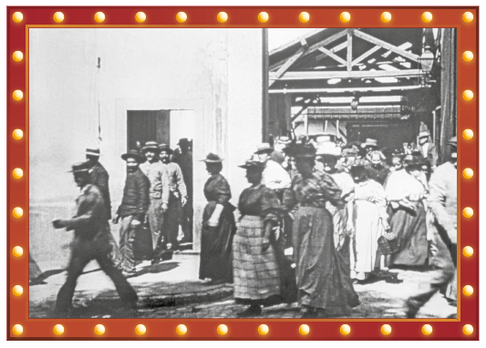 Imagem: Fotografia em preto e branco. Na rua, homens de terno e chapéu e mulheres de longos vestidos e chapéu saem de uma grande construção.  Fim da imagem.