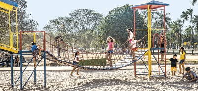 Imagem: Fotografia. Em uma área aberta com chão de areia, muitas crianças brincam em um playground com torres e uma ponte de madeira com cerca de corda.  Fim da imagem.