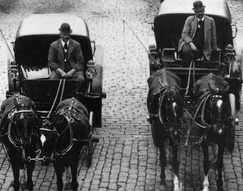Imagem: Fotografia em preto e branco. Dois homens de terno e chapéu coco estão sentados cada um em uma charrete com dois cavalos.   Fim da imagem.
