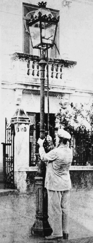 Imagem: Fotografia em preto e branco. Um homem uniformizado com calça, camisa e boina está diante de um poste e manipula uma haste ligada à lamparina.   Fim da imagem.
