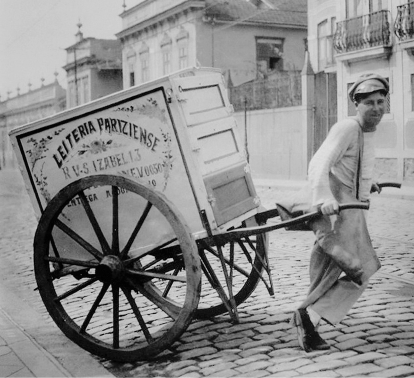 Imagem: Fotografia em preto e branco. Um homem uniformizado com camisa, calça, sapato e boina puxa um carrinho acoplado a uma caixa com a inscrição “LEITERIA PARIZIENSE” em uma rua de paralelepípedos.   Fim da imagem.