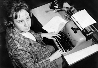 Imagem: Fotografia em preto e branco. Uma mulher de camisa xadrez está sentada diante de uma máquina de escrever sobre uma mesa.  Fim da imagem.