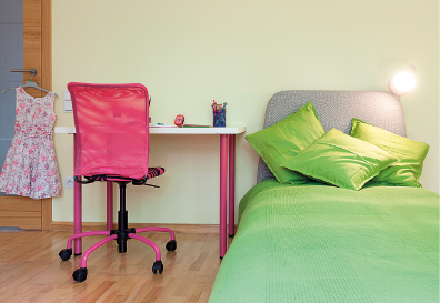 Imagem: Fotografia. Interior de um quarto que apresenta uma cama de solteiro com almofadas, uma luminária ao lado da cabeceira e uma escrivaninha com uma cadeira. Na maçaneta da porta, há um cabide com um vestido pendurado.  Fim da imagem.