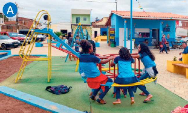 Imagem: A. Fotografia. Em uma praça, crianças uniformizadas brincam no gira-gira de um parquinho. Ao fundo está o escorregador, bancos e uma construção com balcão. Fim da imagem.