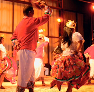 Imagem: Fotografia. Destaque de um grupo que dança em um palco. O homem usa chapéu, camisa vermelha e calça branca e a mulher usa pequeno chapéu, camisa branca e saia de chita.  Fim da imagem.