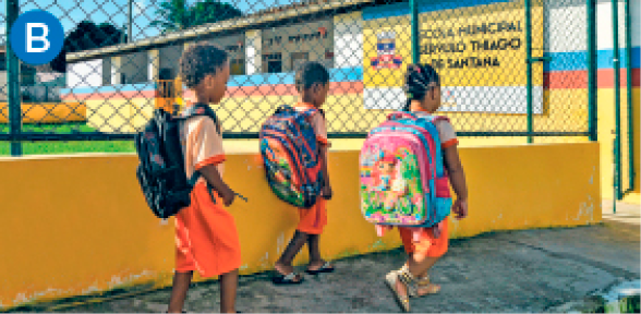Imagem: B. Fotografia. Três crianças uniformizadas caminham com mochilas nas costas na calçada de uma escola com muro baixo e cerca alta.  Fim da imagem.