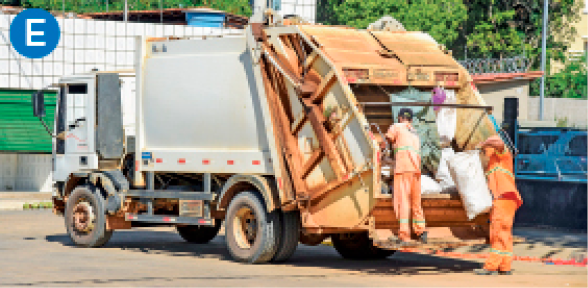 Imagem: E. Fotografia. Um caminhão com caçamba onde estão dois trabalhadores uniformizados. Um deles está sobre o veículo e o outro está no chão segura um saco de lixo. Fim da imagem.