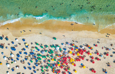 Imagem: Fotografia. Vista aérea da beira-mar com muitos guarda-sóis coloridos abertos na areia e banhistas no mar. Fim da imagem.