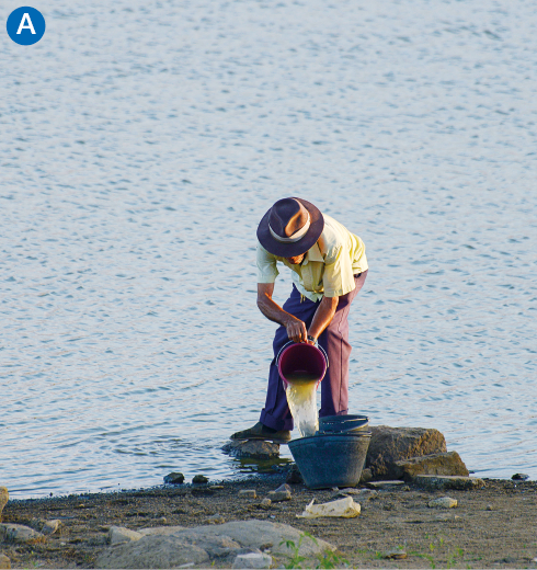 Imagem: Fotografia. Um homem de camisa, calça e chapéu está de pé à beira de um açude. Ele despeja água de um pote em um balde. Fim da imagem.