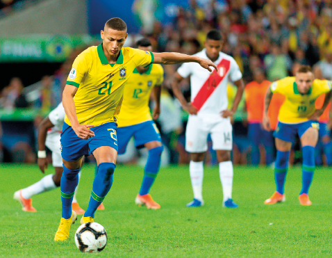 Imagem: Fotografia. Destaque de um campo de futebol onde homens da seleção brasileira jogam. Eles usam camiseta amarela, shorts azuis e meião azul e chuteira. Um jogador do Brasil está em destaque com a bola, os demais observam ao fundo.  Fim da imagem.