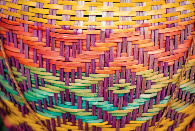 Imagem: Fotografia. Destaque de uma superfície de um cesto com palha colorida entrelaçada formando diferentes grafismos. Fim da imagem.