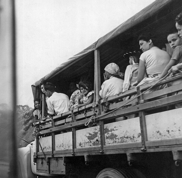 Imagem: Fotografia em preto e branco. Destaque da carroceria de um caminhão com a lateral aberta e o teto coberto e onde estão muitas pessoas sentadas, entre elas mulheres, crianças e alguns homens.  Fim da imagem.