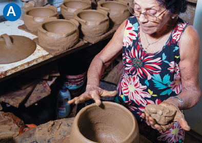 Imagem: A. Fotografia. Em uma sala, uma senhora de óculos está sentada e constrói um vaso com barro. Ao lado, uma mesa com muitos potes moldados. Fim da imagem.