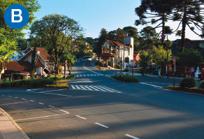 Imagem: B. Fotografia. Destaque de uma via asfaltada pintada com sinalização de trânsito. Na região, há casas e árvores.  Fim da imagem.