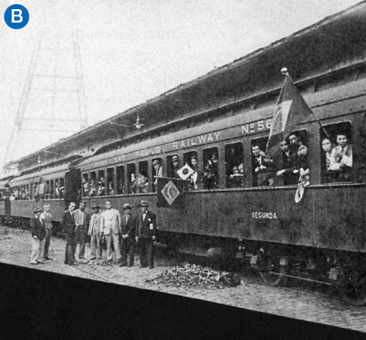 Imagem: B. Fotografia. Destaque de um trem lotado de pessoas e algumas aparecem nas janelas, onde estão bandeiras do Brasil e do Japão hasteadas. Do lado de fora, à frente do transporte, está um grupo de oito homens de terno, alguns de chapéu, parados posando para foto.  Fim da imagem.