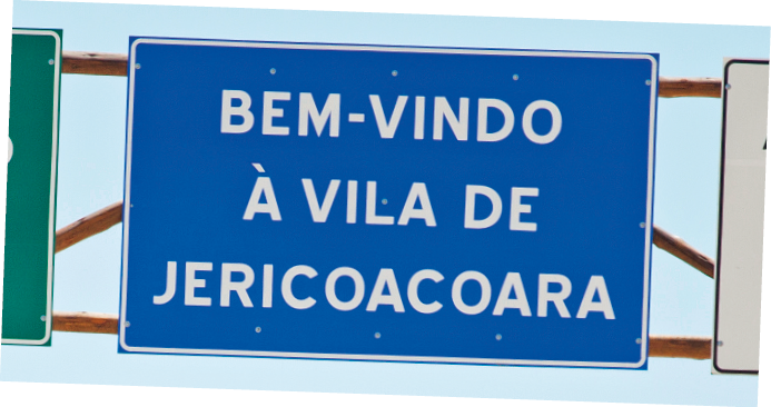 Imagem: Fotografia. Uma placa grande em cor azul onde está escrito em letras com caixa alta “BEM-VINDO À VILA DE JERICOACOARA”.  Fim da imagem.