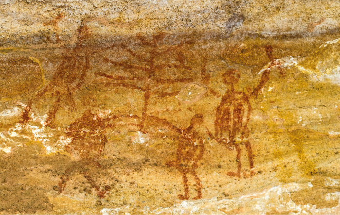 Imagem: Fotografia. Em uma superfície rochosa amarelada, pintura de quatro seres humanos de pé ao redor de uma figura comprida com muitos filamentos. Fim da imagem.