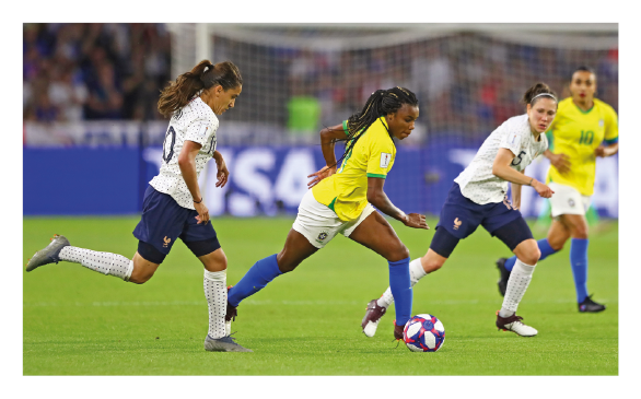 Imagem: Fotografia. Destaque de um campo de futebol onde mulheres da seleção brasileira jogam. Elas usam camiseta amarela, shorts brancos, meião azul e chuteira. Há uma jogadora do Brasil com a bola entre duas jogadoras do time adversário.  Fim da imagem.