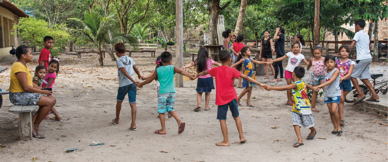 Imagem: Fotografia. Plano aberto que apresenta um local com chão de terra, pequenas casas, bancos e árvores.  Em destaque, estão muitas crianças. Elas fazem uma roda de mãos dadas. Ao redor, algumas outras observam e há poucos adultos, que conversam. No canto direito, há uma bicicleta no chão. Fim da imagem.