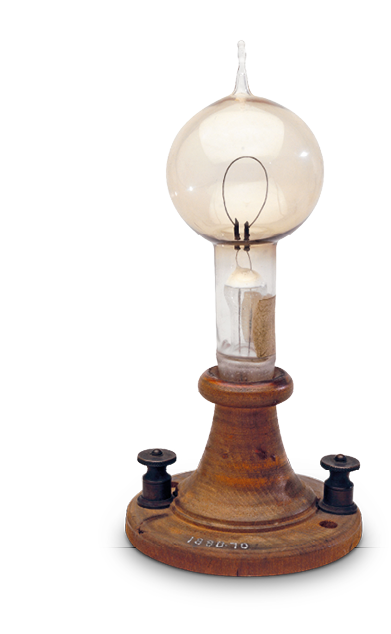 Imagem: Fotografia. Uma lâmpada com base cilíndrica e corpo redondo com uma pequena saliência fina na parte superior. Internamente apresenta um filamento com formato oval. A lâmpada está uma base circula de madeira.  Fim da imagem.