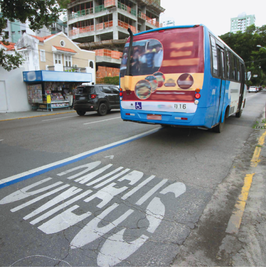 Imagem: Fotografia. Destaque de uma via onde trafega um ônibus e no asfalto está escrito “ÔNIBUS” em uma faixa exclusiva. Ao fundo, um carro, casa e um prédio em construção.  Fim da imagem.