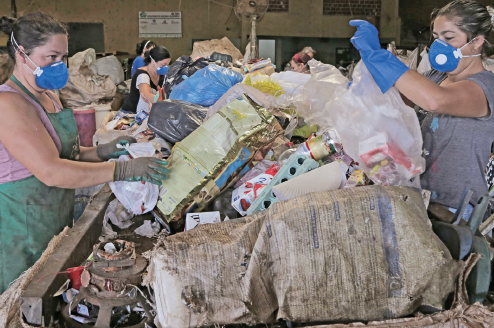Imagem: Fotografia. Destaque de uma esteira onde há muitos materiais recicláveis amontoados e que são manipulados por mulheres que usam luvas e máscaras.  Fim da imagem.