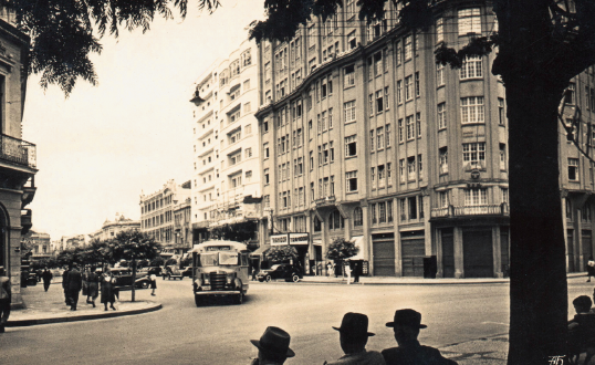 Imagem: Fotografia em preto e branco. Em primeiro plano, homens de chapéu sentados ao lado de uma árvore. Em segundo plano, uma via pela qual trafega um veículo e pessoas caminham na calçada. Na calçada, destaque de um alto e amplo edifício com muitas janelas.  Fim da imagem.